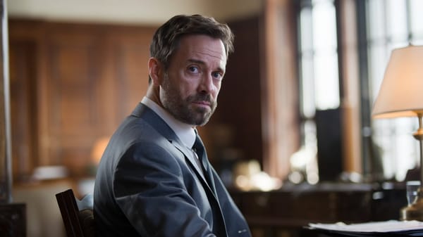 A cinematic shot of a middle-aged man with a beard sitting in a room.