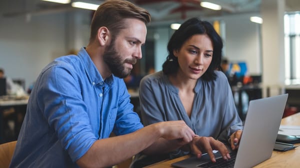 A photo of a man with a beard and a woman with dark hair are working on a laptop in an office setting.