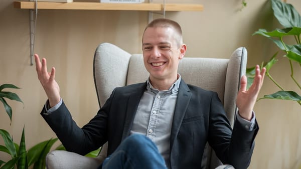 A photo of a man with short hair, wearing a gray shirt and a black blazer, sitting in a comfortable chair.